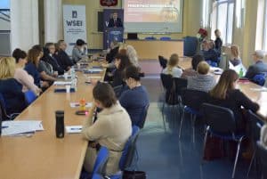 Grupa osób zasiadająca przy długich stołach i patrząca na audytorium oraz osobę stojącą przy mównicy. Za mównicą wisi na ścianie ekran projektowy na którym jest wyświetlana grafika z tytułem “pokonywać niepokonane”