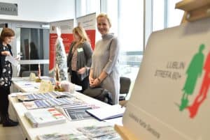 Dwie kobiety stojące przy długim stole na którym są papierowe broszury i ulotki. Jedna z kobiet uśmiecha się do zdjęcia.