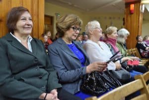 Obiektyw skierowany jest na widownię. Na pierwszym planie na krzesłach siedzi pięć kobiet, druga od lewej na kolanach trzyma torebkę a w dłoniach broszurę. W tle znajdują się inne osoby z widowni, dwie drewniane kolumny oraz krzesła.