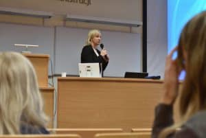 Na scenie auli wykładowej stoi Pani z mikrofonem trzymanym oburącz. Po obu stronach zdjęcia widać włosy kobiet siedzących na auli.