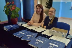Dwie młode uśmiechające się do zdjęcia kobiety siedzą przy stole z teczkami z logiem WSEI i innymi materiałami papierowymi.