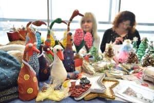 Kobiety siedzące przy stole z rękodziełami. Są łabędzie robione z różnych materiałów o różnych wzorach. W tle choinki z wosku.