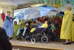 Na scenie znajduje się dwanaście osób, cztery z nich to dzieci z niepełnosprawnością na wózkach. Inne osoby stoją w półokręgu obok nich w kolorowych – niebieskie, żółte, pomarańczowe – tunikach i trzymają nad nimi biało – niebieski materiał.