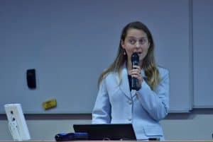 Pani w jasnym żakiecie trzyma w prawym ręku mikrofon. W tle jest biała tablica.