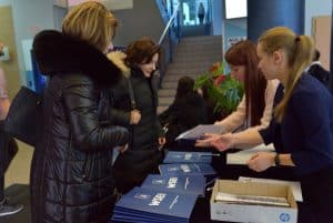 Dwie młode kobiety stoją przy stole i rozdają innym osobom, które podeszły do stołu teczki z logiem WSEI.