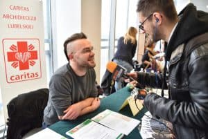 Mężczyzna siedzący przy stole z rękoma założonymi na siebie uśmiecha się do mężczyzny, który się nachyla do niego. W tle baner reklamowy z grafiką caritasu na której jest czerwony plus w środku którego jest napis cartias układający się w kształt serca.