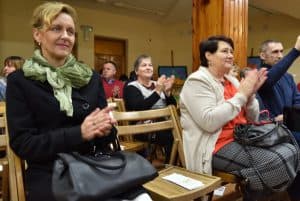 Obiektyw skierowany jest na widownię. Na pierwszym planie w środku widzimy puste krzesło a na nim broszurę, po prawej i lewej stronie krzesła siedzą dwie kobiety. W tle znajdują się inne osoby z widowni, drewniana kolumna oraz drewniane drzwi.