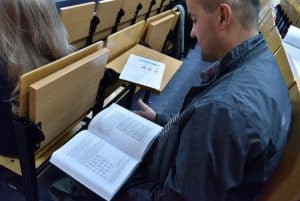 Pan siedzący w auli trzyma na kolanach książkę i ma wzrok skierowany na nią. Przed nim w następnym rzędzie siedzi kobieta z długimi włosami.