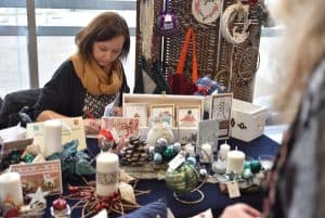 Kobieta siedzi przy stole z rękodziełami i wyrobami manualnymi. Trzyma w dłoni długopis. Dłoń ma położoną na kartce