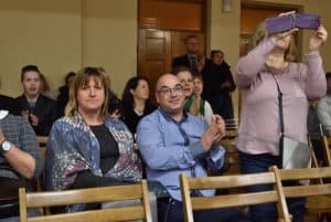 Obiektyw skierowany jest na widownię. Na pierwszym planie w środku widzimy oparcia trzech pustych krzeseł. Z tyłu od prawej siedzi kobieta i mężczyzna, po lewej stoi kobieta trzymająca telefon przed twarzą.