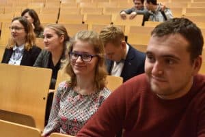 Młodzi ludzie siedzą na auli. Na pierwszym planie jest kobieta i mężczyzna którzy się uśmiechają