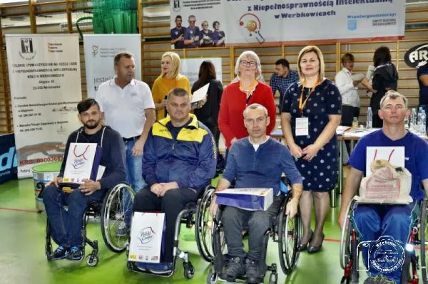 Ludzie siedzący na wózkach inwalidzkich na sali gimnastycznej. Na kolanach trzymają torby prezentowe. Za nimi stoją ludzie.