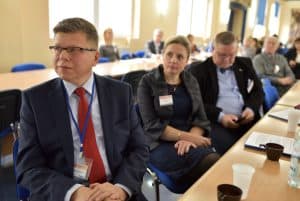 Trzech mężczyzn i jedna kobieta siedzą przy długim stole i patrzą na wprost do zdjęcia. W tle za nimi inne osoby siedzące przy tym samym stole. Na stole znajdują się materiały papierowe, plastikowy kubeczek, filiżanka i butelka z wodą mineralną.