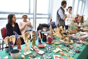 Ludzie siedzący i stojący za stołem z wyrobami manualnymi i rękodziełami. Na stole są ręcznie robione aniołki, renifery i choinki.