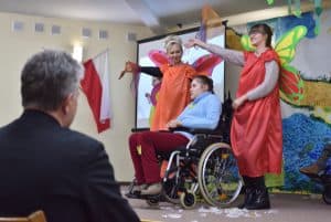 Na scenie znajdują się trzy osoby, dwie kobiety w kolorowych tunikach ze skrzydłami stoją po lewej i prawej stronie dziewczyny z niepełnosprawnością przebywającej na wózku. Na pierwszym planie widać mężczyznę odwróconego plecami do obiektywu.