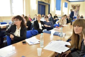 Grupa osób siedząca przy długim stole i patrząca na wprost. Na pierwszym planie zdjęcia znajdują się dwie kobiety - jedna patrzy przed siebie a druga patrzy wprost do zdjęcia.