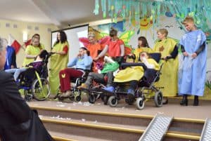 Na scenie znajduje się trzynaście osób w tym cztery osoby z niepełnosprawnością przebywające na wózkach. Dziewięć osób stojących za wózkami ma na sobie kolorowe tuniki: żółte, niebieskie, pomarańczowe. Na pierwszym planie po prawej stronie widać rampę do wjazdu wózków na scenę.