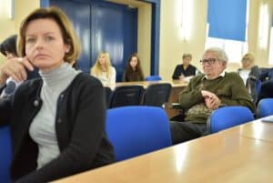 Kobieta siedząca przy stole i patrząca na wprost do zdjęcia. Głowę opiera o rękę założoną na krzesło na którym siedzi.