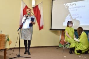 Po lewej stronie za mikrofonem stoi kobieta, prawą rękę trzyma na mikrofonie a w lewej ma niebieską teczkę. Za nią znajdują się dwie biało – czerwone flagi. Po prawej na krześle siedzi dziewczynka w żółtej tunice, obok niej kuca kobieta w takim samym stroju.