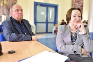 Kobieta siedząca przy końcu stołu razem uśmiecha się i podpiera swoją brodę o założona ręka na ręce dłoń. Za nią siedzący mężczyzna i patrzący przed siebie, na wprost do zdjęcia.