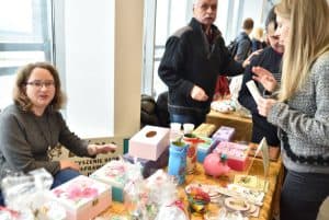 Kobieta siedzi przy stole z rękodziełami i wyrobami manualnymi. W tle inni ludzie oraz kobieta która stoi przy tym stanowisku i w dłoni trzyma zgiętą na pół kartkę.