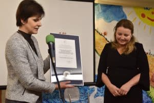 Na pierwszym planie znajdują się dwie kobiety, pani po lewej stronie stoi za mikrofonem a w obu rękach trzyma dokument w ramce. W tle znajduje się ekran do rzutnika oraz kolorowa ściana.