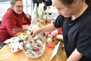 Kobieta siedzi przy stole z rękodziełami. Naprzeciwko niej kobieta wkłada dłoń do przezroczystej szklanej kuli w rękodziełami.