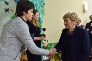 Na pierwszym planie stoją dwie kobiety, podają sobie ręce w geście gratulacyjnym, ta po lewej trzyma w drugiej ręce niebieski dokument. Nieco dalej stoi kolejna kobieta trzymając w obu rękach różę.