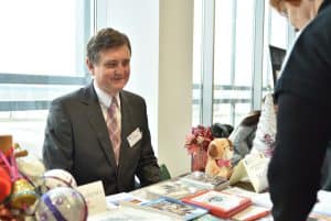 mężczyzna ubrany w garnitur siedzi przy stole zastawionym rękodziełami. Uśmiecha się.