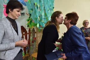 Na pierwszym planie znajdują się trzy kobiety. Druga i trzecia – licząc od lewej – podają sobie ręce a ich twarze skierowane są ku sobie. W tle znajduje się scenografia przedstawiająca drzewo z papieru i bibuły.