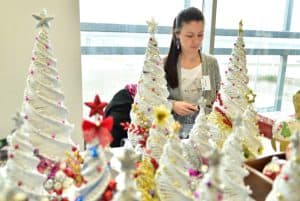 Kobieta siedzi przy stole z rękodziełami. Wśród wyrobów manualnych głównie znajdują się białe choinki.