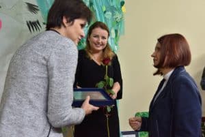 Na pierwszym planie stoją dwie kobiety, pani po lewej trzyma w rękach ramkę. Nieco dalej stoi kolejna kobieta trzymając w obu rękach różę.