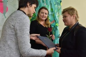 Na pierwszym planie profilem stoją dwie kobiety, trzymają wspólnie w rękach niebieską teczkę. Nieco dalej stoi kolejna kobieta trzymając w obu rękach różę.