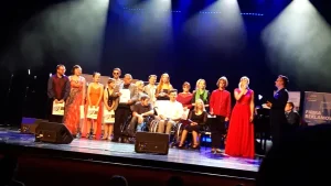 Na scenie na sali jest duża grupa ludzi. Część z nich siedzi na wózkach inwalidzkich, a część stoi. Na tych ludzi skierowane jest całe oświetlenie sceny.