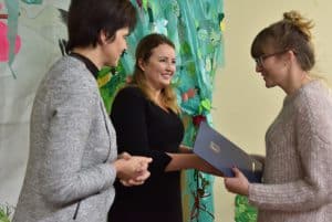 Na pierwszym planie znajdują się trzy kobiety. Druga i trzecia – licząc od lewej – podają sobie ręce. Pani w środku trzyma różę, pani po prawej trzyma też niebieską teczkę.