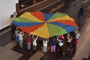 Dzieci trzymające dużą kolorową chustę animacyjna w kształcie koła. W oddali mężczyzna trzymający w dłoniach aparat fotograficzny.