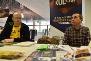 Dwoje mężczyzn siedzących przy stole z broszurami i ulotkami. W tle baner z napisem KUL.