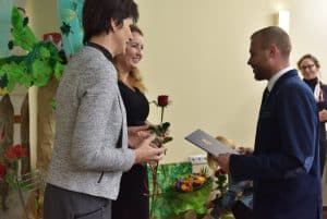 Na pierwszym planie znajdują się dwie kobiety i mężczyzna. Pani w środku trzyma różę, a mężczyzna po prawej trzyma w obu rękach niebieską teczkę.