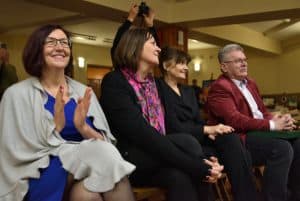 Obiektyw skierowany jest na widownię. Na pierwszym planie znajdują się trzy kobiety i mężczyzna, który na swoich kolanach trzyma zielone pudełko. Pani po prawej trzyma ręce w geście bicia brawa.