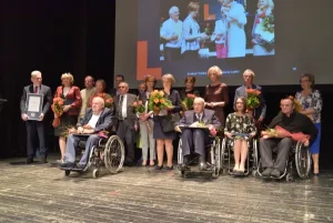 Na scenie jest duża grupa ludzi. Kilka z nich siedzi na wózkach inwalidzkich. W tle jest ekran na którym jest wyświetlone zdjęcie. Kilka osób trzyma w rękach kwiaty.