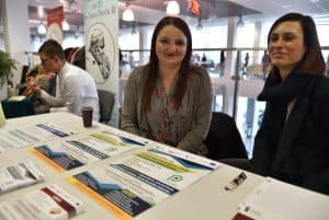 Dwie kobiety siedzące przy stole z plakatami. Uśmiechają się do zdjęcia.