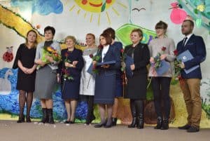 Na scenie stoi 9 osób w tym jeden mężczyzna i osiem kobiet. Sześć osób trzyma w ręce czerwoną różę, druga od lewej ma w rękach duży bukiet. W tle znajduje się kolorowa ściana z między innymi: łabędziem, biedronką, słońcem, chmurami.