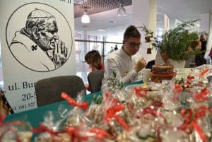 Młody mężczyzna siedzący przy stole z rękodziełami zapakowanymi w przeźroczyste celofany i przewiązane u góry czerwoną kokardą.