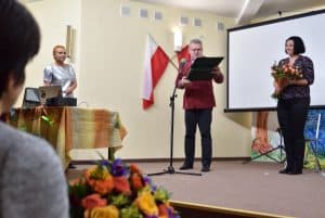 Na scenie znajdują się trzy osoby. Na środku za mikrofonem stoi mężczyzna z otwartym zielonym pudełkiem. Po prawej stoi kobieta trzymająca bukiet kwiatów, za nią znajduje się ekran do wyświetlania prezentacji multimedialnych. Kobieta po lewej stoi za stołem na którym znajduje się laptop.