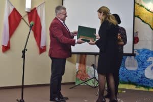 Na pierwszym planie mężczyzna i kobieta trzymają otwarte pudełko z dyplomem. Nieco dalej stoi pani trzymająca bukiet. W tle znajduje się ekran do wyświetlania prezentacji oraz kolorowa ściana z dwoma łabędziami i biedronką.