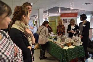 Ludzie stojący wokół stołu z rękodziełami manualnymi. Na stole wiklinowe koszyki i różne kartki.