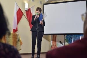 Na pierwszym planie znajdują się tyłem do obiektywu ludzie, ich sylwetki nie są w pełni widoczne. Na scenie za mikrofonem stoi kobieta trzymająca w prawej ręce mikrofon a w lewej teczkę i pakunek w folii. W tle widać ekran do wyświetlania prezentacji.