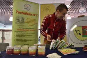 Młody mężczyzna ubrany w dres stoi przy stole z rękodziełami. W tle baner reklamowy Technikum nr 1.