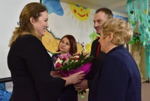 Cztery osoby stoją twarzami do siebie, kobieta po lewej widoczna z profilu wraz z mężczyzną po prawej trzymają bukiet kwiatów. W tle znajduje się kolorowa ściana z rysunkiem chmur, słońca oraz drogi.