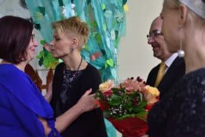 Dwie kobiety po lewej stoją twarzami do siebie, ta po lewej trzyma ręce na ramionach drugiej. Po prawej na pierwszym planie znajduje się rozmazana sylwetka kobiety z profilu, nieco dalej stoi mężczyzna z bukietem kwiatów.